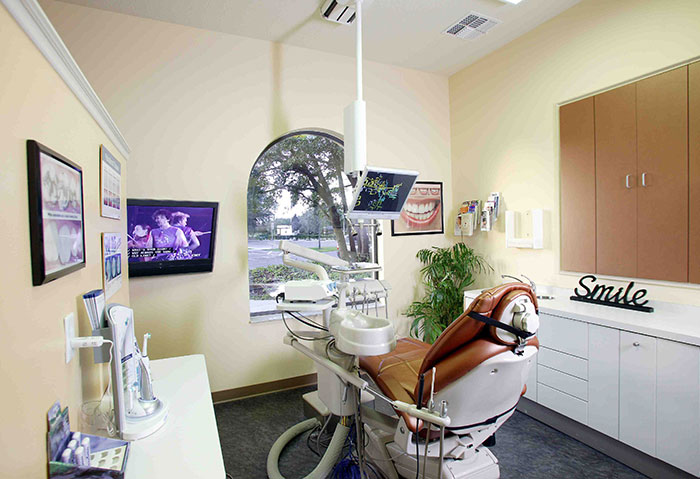The image shows a modern dental office interior with a large window, a reception area, and a well-equipped dental chair.